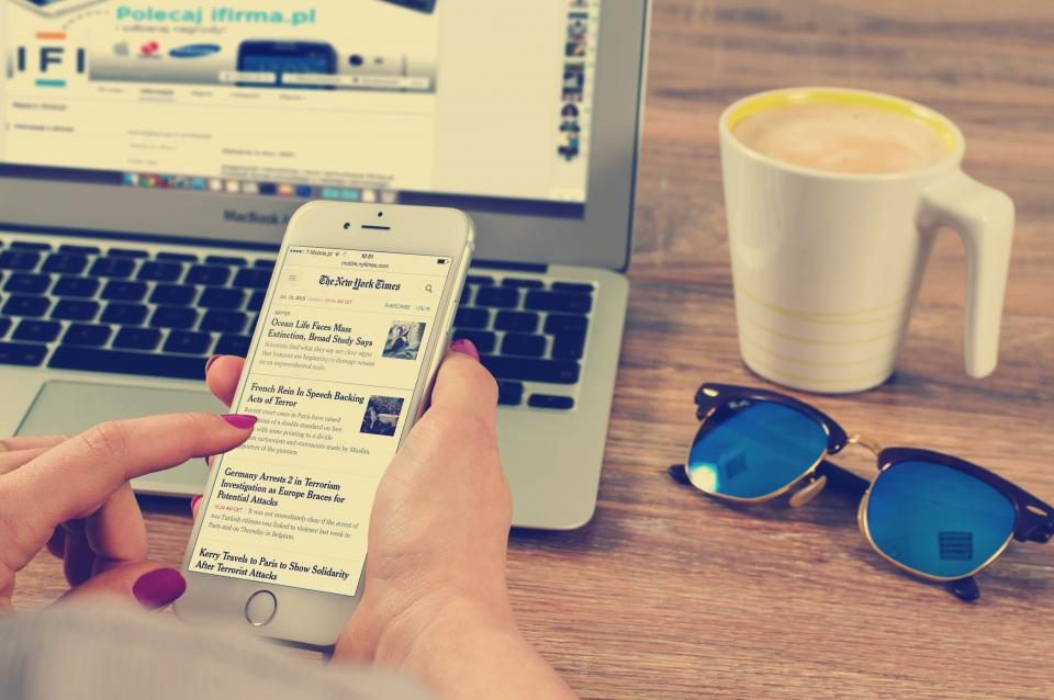 Pessoa rolando um feed no celular em frente ao notebook, com café e óculos de sol na mesa