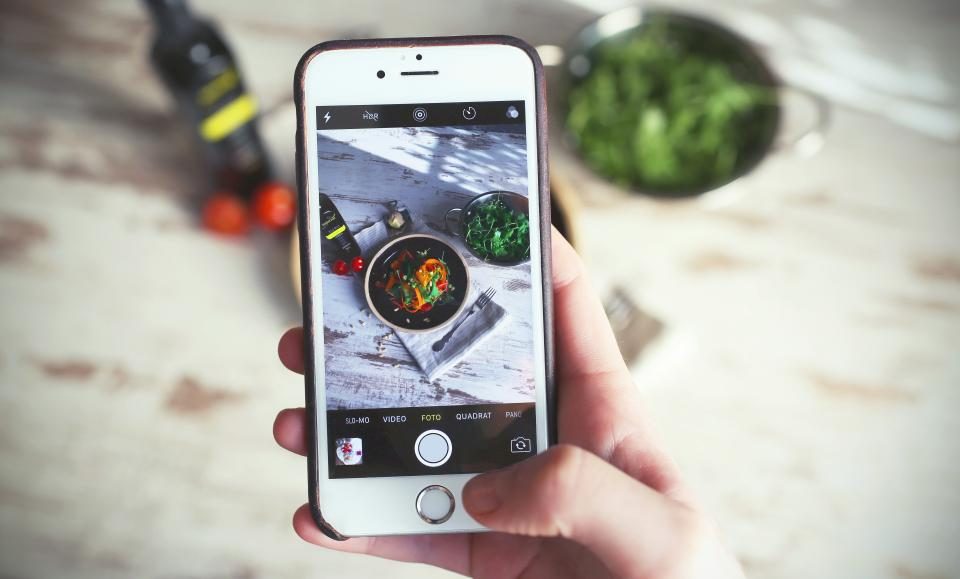 Mão segurando o celular enquanto fotografa um prato de comida sobre a mesa