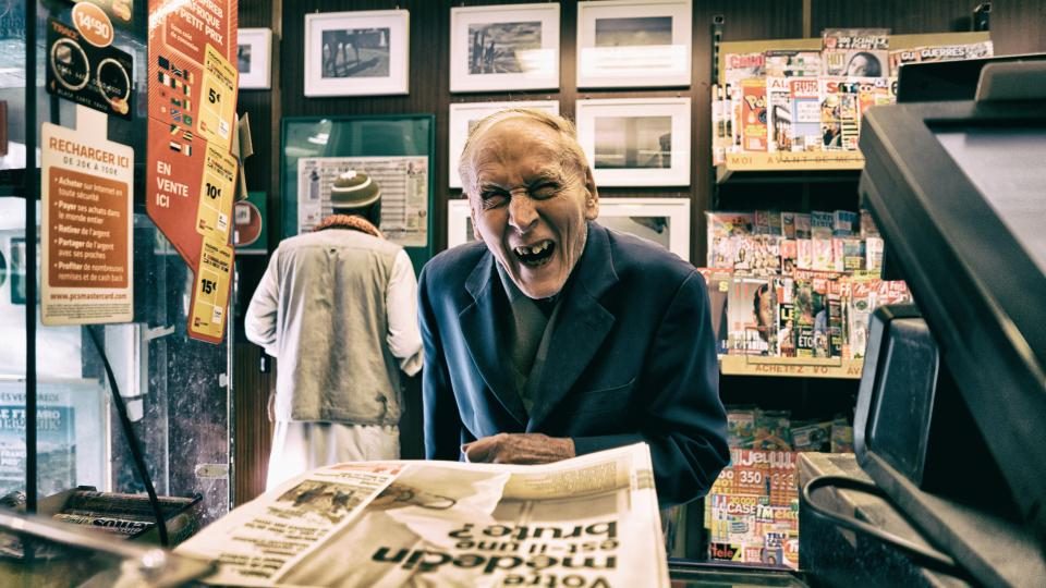 Comerciante idoso sorrindo atrás do balcão de uma banca cheia de quadros e jornais