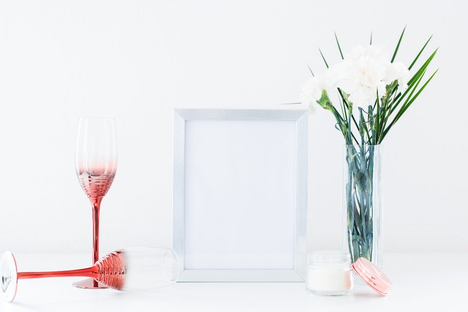 Composição de objetos sobre fundo branco, com um porta-retratos, um vaso de flores e uma taça