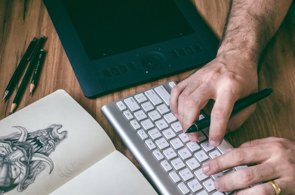 Designer desenhando em um caderno ao lado de uma mesa digitalizadora e de um teclado