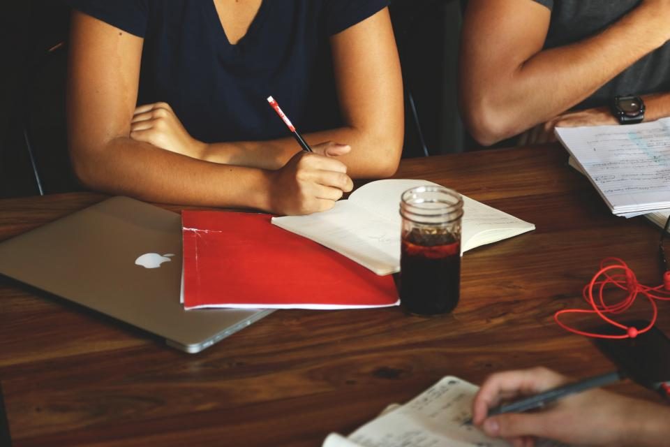 Pessoas à mesa fazendo anotações, com um caderno vermelho e um notebook