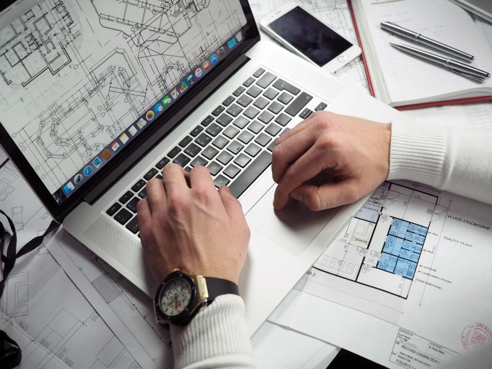 Pessoa trabalhando no notebook ao lado de folhas com esboços de layout de site