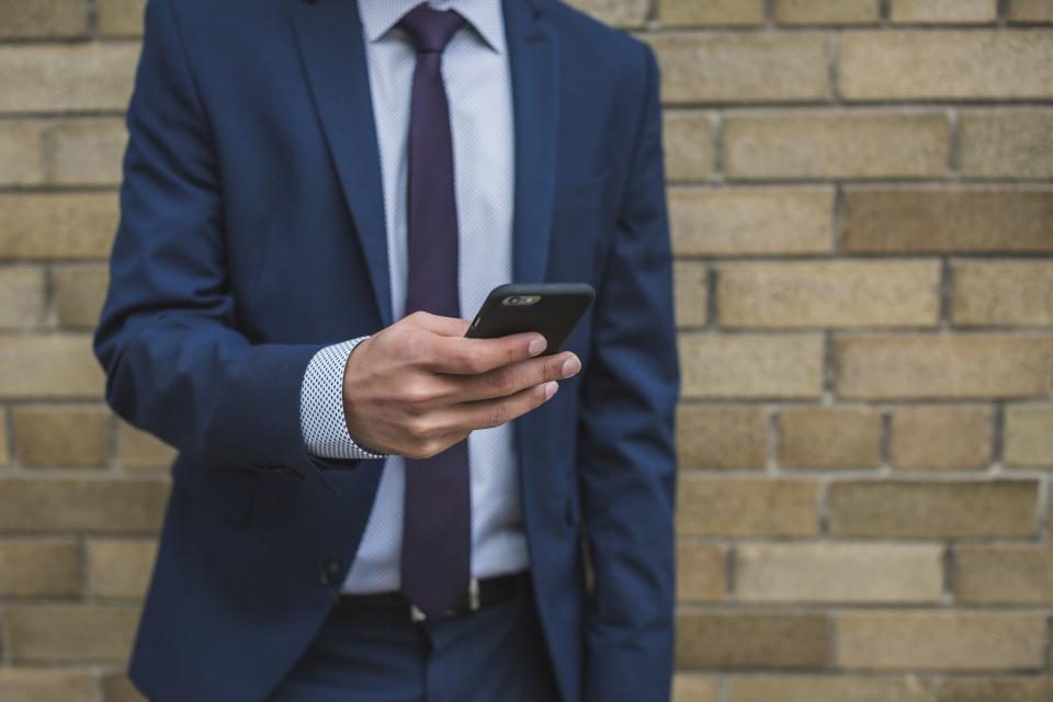 Homem de terno usando o celular, encostado numa parede de tijolos
