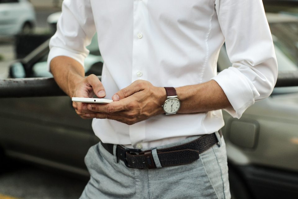 Homem de camisa branca segurando e usando o celular, encostado num carro