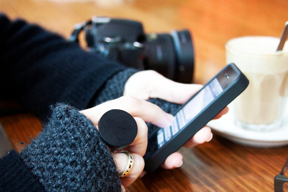 Mão segurando o celular ao lado de uma câmera fotográfica e de uma xícara de café
