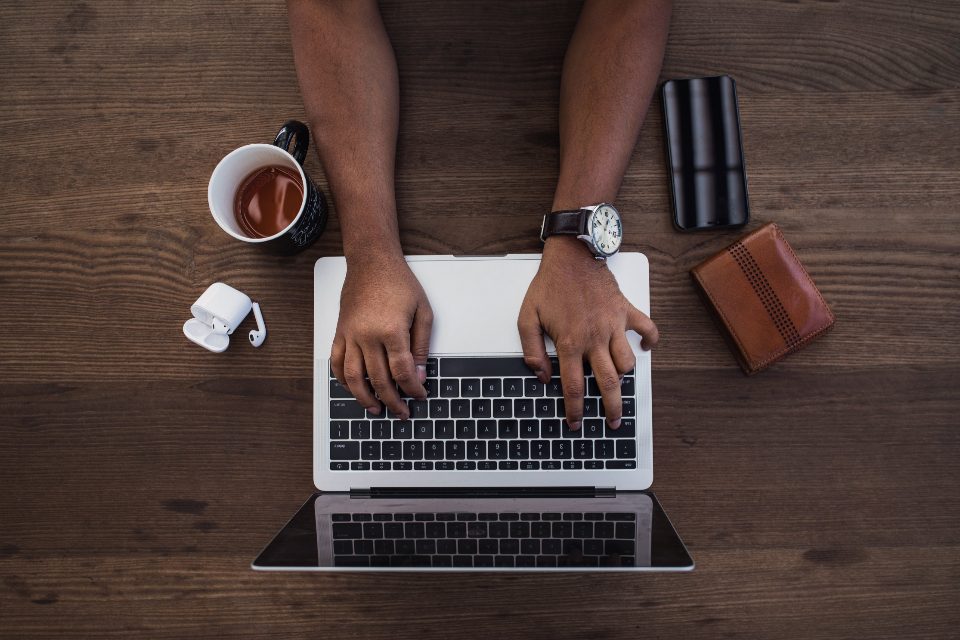 Vista de cima de mãos digitando num notebook, com uma xícara de café e uma carteira na mesa