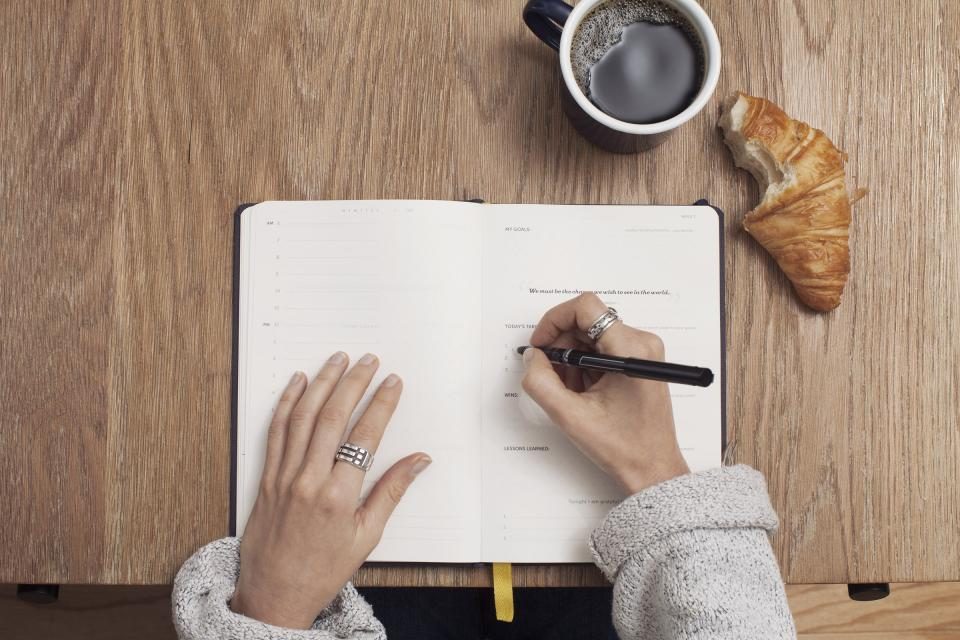 Mãos escrevendo em um caderno sobre a mesa, ao lado de um café e um croissant