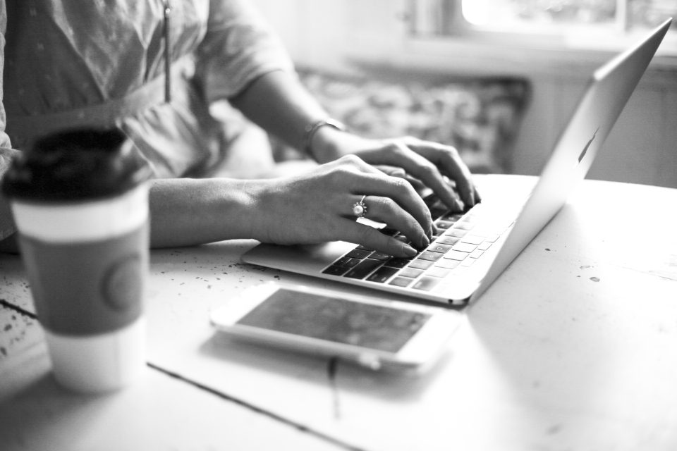 Foto em preto e branco de mãos digitando num notebook, com um celular e um copo de café na mesa
