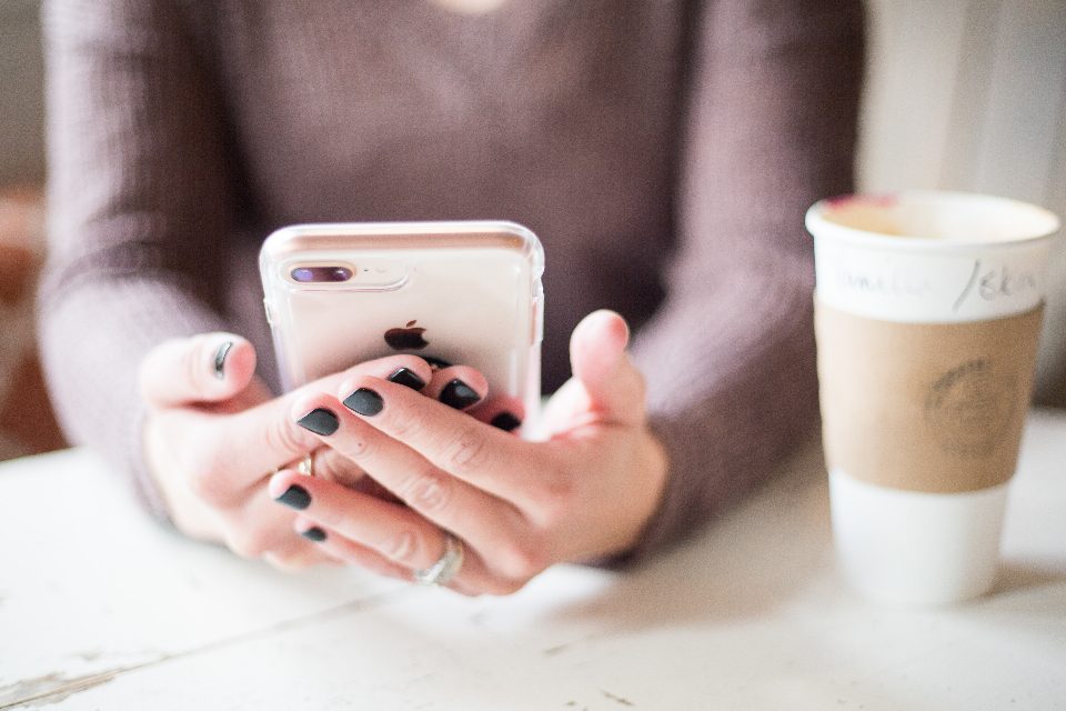 Mãos de mulher segurando um celular sobre a mesa, ao lado de um copo de café