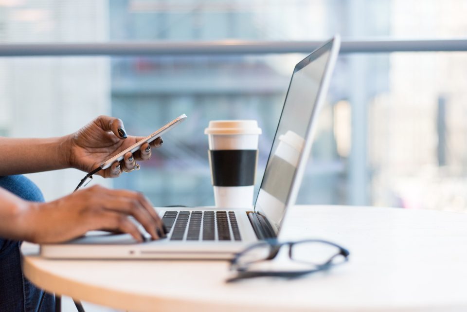 Mãos usando o celular ao lado de um notebook aberto, com um copo de café na mesa