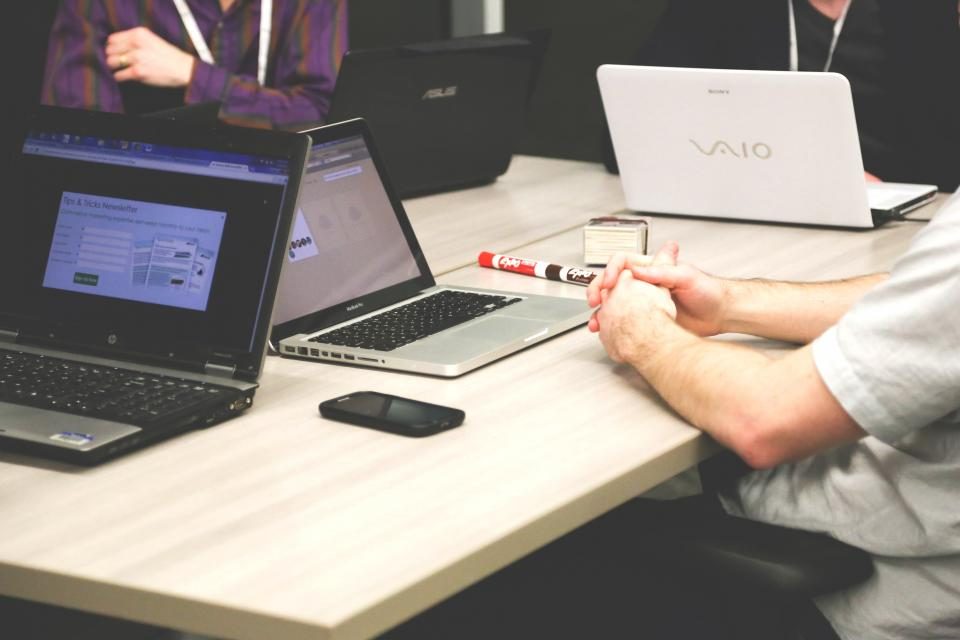 Mesa de trabalho com dois notebooks abertos e uma pessoa fazendo anotações