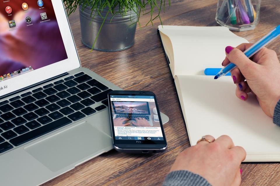 Mesa com notebook, celular e uma pessoa escrevendo num caderno