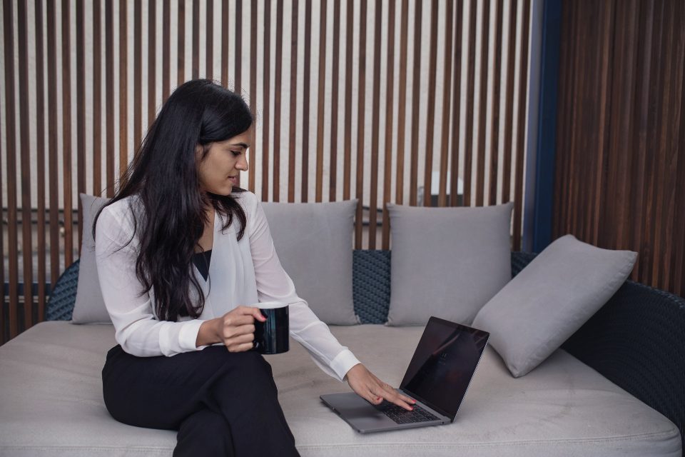 Mulher trabalhando no notebook sentada no sofá, com uma xícara na mão