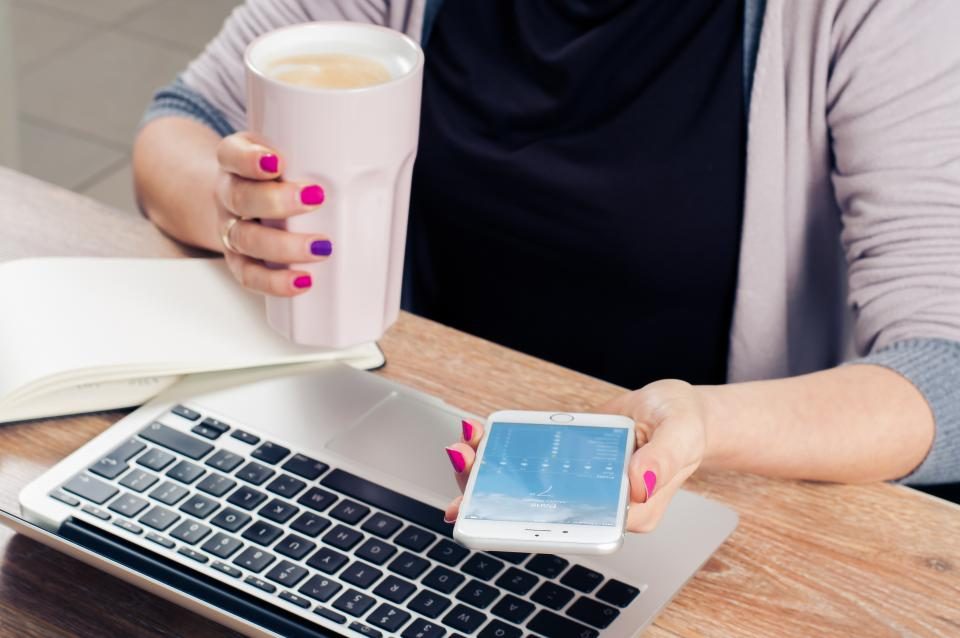 Mulher segurando o celular com uma mão e uma xícara com a outra, perto de um notebook