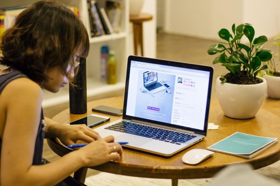 Mulher trabalhando num notebook numa mesa em casa, ao lado de uma planta