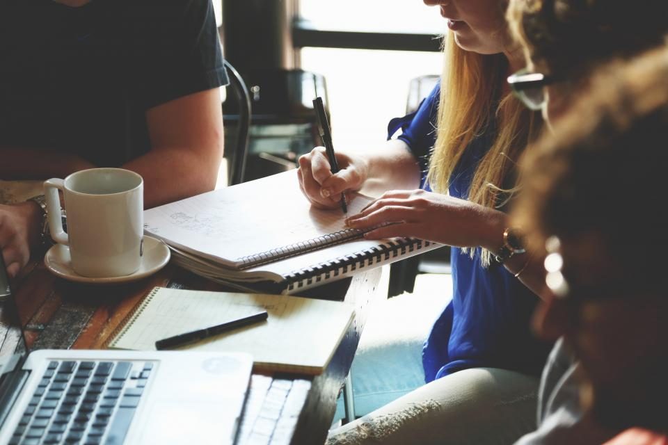 Pessoas reunidas em volta de uma mesa com notebook, cadernos e café, uma delas fazendo anotações